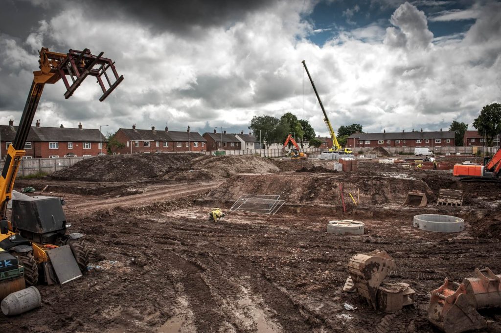 Bouwplaats met graafmachines en huizen op achtergrond.